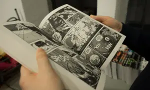 Mani che sfogliano un libro di graphic journalism con scene d'azione e dialoghi in stile fumetto.
