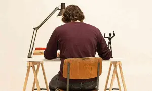 Uomo seduto alla scrivania, illuminato da una lampada, con libri e una statuetta. Il suo **titolo clickbait** è un mistero!