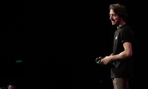 Un uomo con una maglietta nera con la lettera "M" parla sul palco, simile a uno dei tanti blog italiani famosi.
