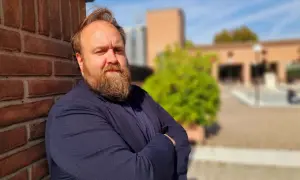 Nicolas Ballario, con barba folta e sguardo intenso, appoggiato a un muro di mattoni rossi, con un blazer blu.