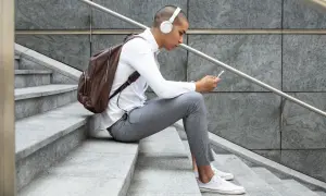 Giovane con cuffie bianche e zaino marrone seduto sulle scale, smartphone in mano, perfetto per scoprire dove ascoltare podcast.