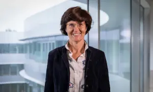 Paola Dubini, sorridente con occhiali e giacca scura, sullo sfondo di un edificio moderno con ampie vetrate.
