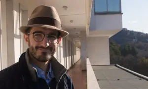 Ruggero Rollini, con cappello e occhiali, sorride su un balcone con vista su un paesaggio collinare.