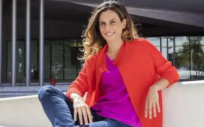 Francesca Bria sorride, seduta all'esterno con un cardigan arancione acceso e una maglia fucsia.