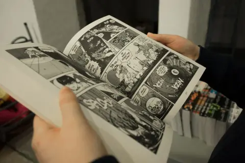 Mani che sfogliano un libro di graphic journalism con scene d'azione e dialoghi in stile fumetto.