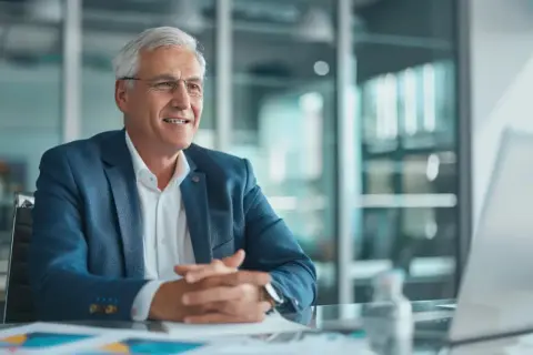 Un uomo anziano con capelli grigi e occhiali, vestito con giacca blu, sorride mentre discute di affari. Potrebbe essere il CFO che spiega i risultati finanziari.