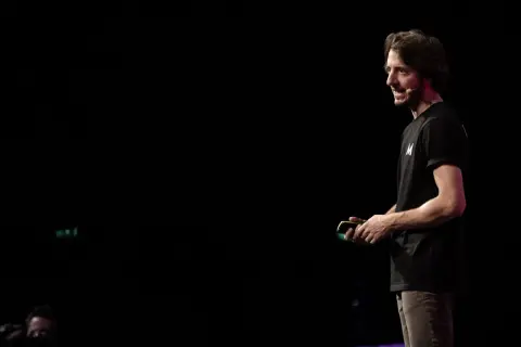 Un uomo con una maglietta nera con la lettera "M" parla sul palco, simile a uno dei tanti blog italiani famosi.