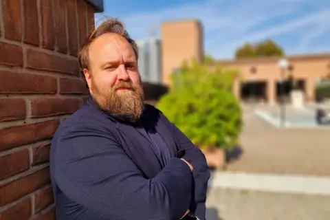 Nicolas Ballario, con barba folta e sguardo intenso, appoggiato a un muro di mattoni rossi, con un blazer blu.
