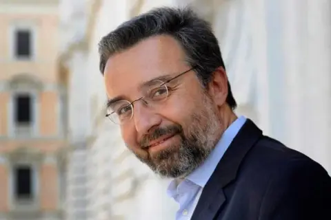 Marco Damilano, giornalista con barba e occhiali, sorride con un blazer scuro.