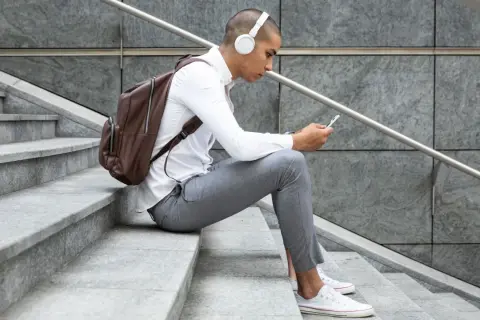 Giovane con cuffie bianche e zaino marrone seduto sulle scale, smartphone in mano, perfetto per scoprire dove ascoltare podcast.