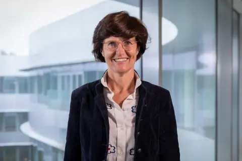 Paola Dubini, sorridente con occhiali e giacca scura, sullo sfondo di un edificio moderno con ampie vetrate.