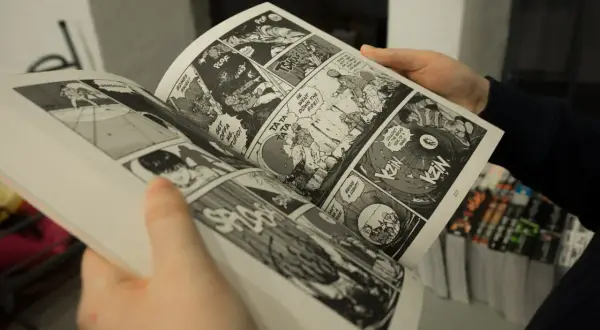 Mani che sfogliano un libro di graphic journalism con scene d'azione e dialoghi in stile fumetto.