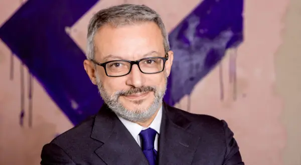Gianluca Foglia, con occhiali e cravatta blu, sorride davanti a uno sfondo astratto viola e rosa.