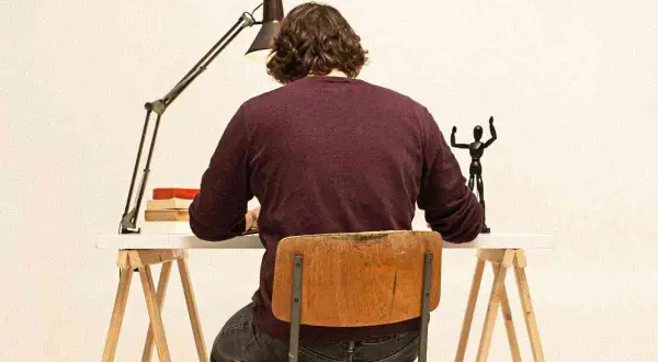 Uomo seduto alla scrivania, illuminato da una lampada, con libri e una statuetta. Il suo **titolo clickbait** è un mistero!