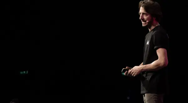 Un uomo con una maglietta nera con la lettera "M" parla sul palco, simile a uno dei tanti blog italiani famosi.