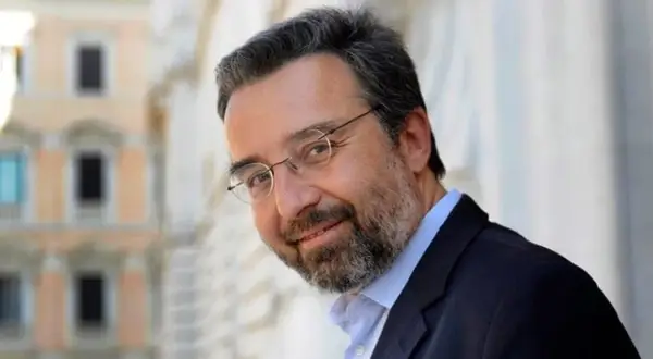 Marco Damilano, giornalista con barba e occhiali, sorride con un blazer scuro.