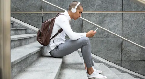 Giovane con cuffie bianche e zaino marrone seduto sulle scale, smartphone in mano, perfetto per scoprire dove ascoltare podcast.