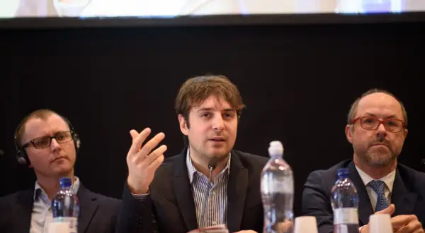 Beniamino Pagliaro parla a un microfono, con altri due uomini seduti al suo fianco.