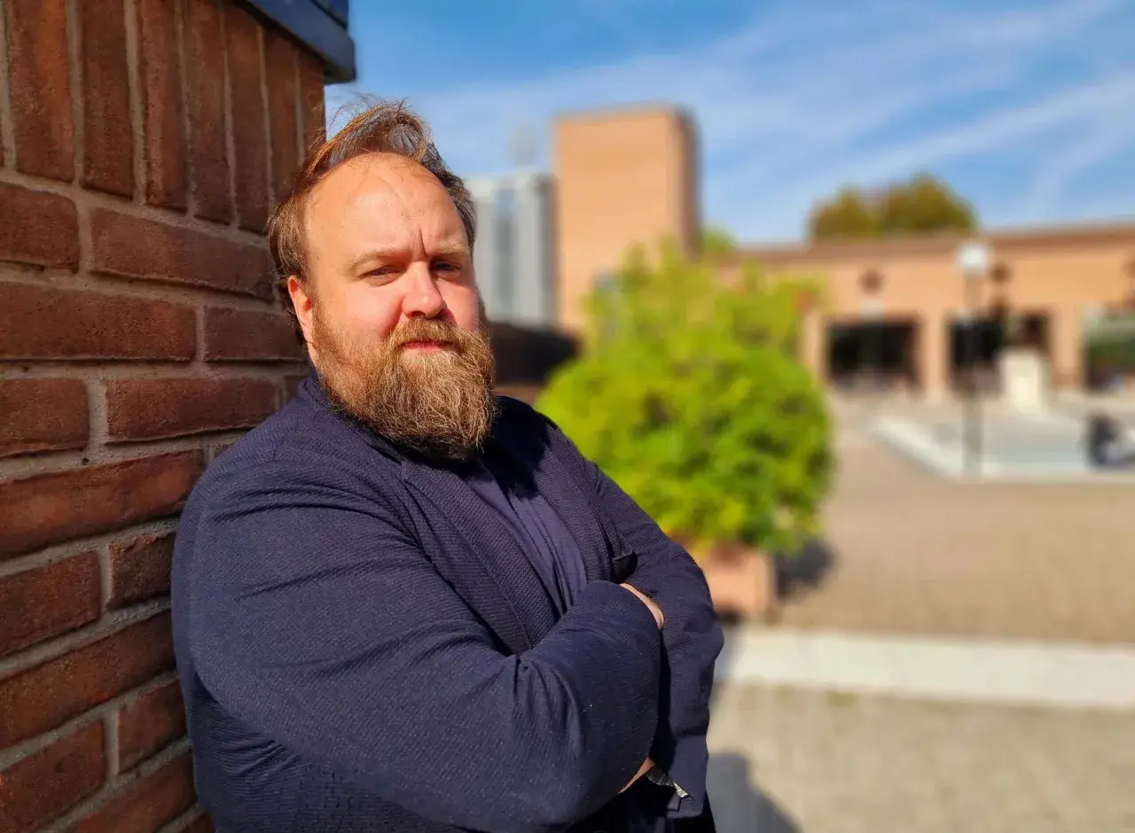 Nicolas Ballario, con barba folta e sguardo intenso, appoggiato a un muro di mattoni rossi, con un blazer blu.