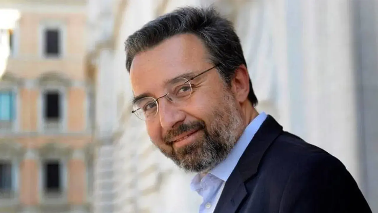 Marco Damilano, giornalista con barba e occhiali, sorride con un blazer scuro.