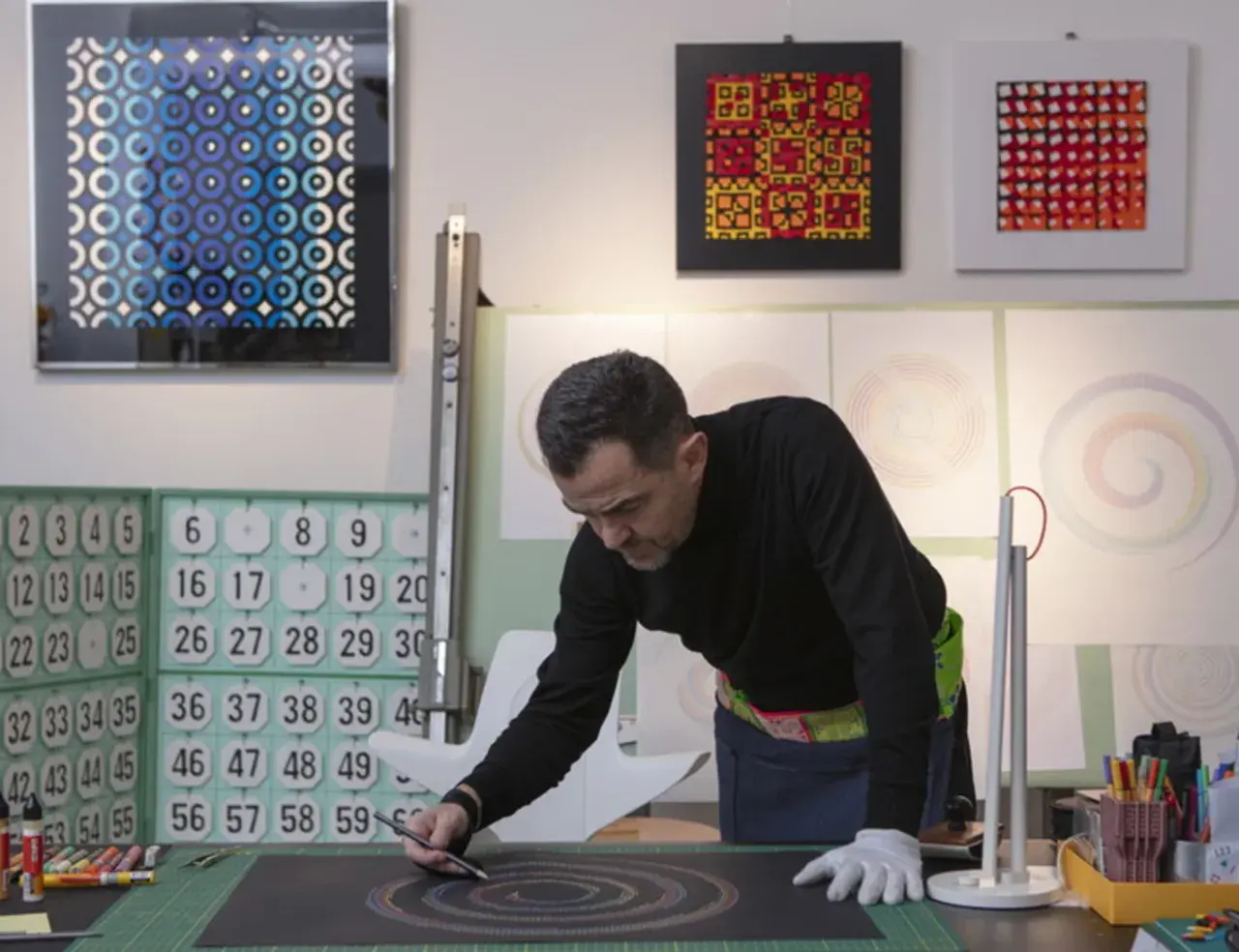 Adriano Attus, artista, disegna cerchi colorati su una tela nera. Sullo sfondo, opere astratte e numeri.