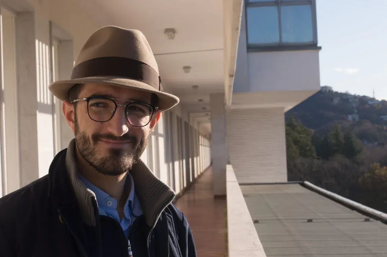 Ruggero Rollini, con cappello e occhiali, sorride su un balcone con vista su un paesaggio collinare.