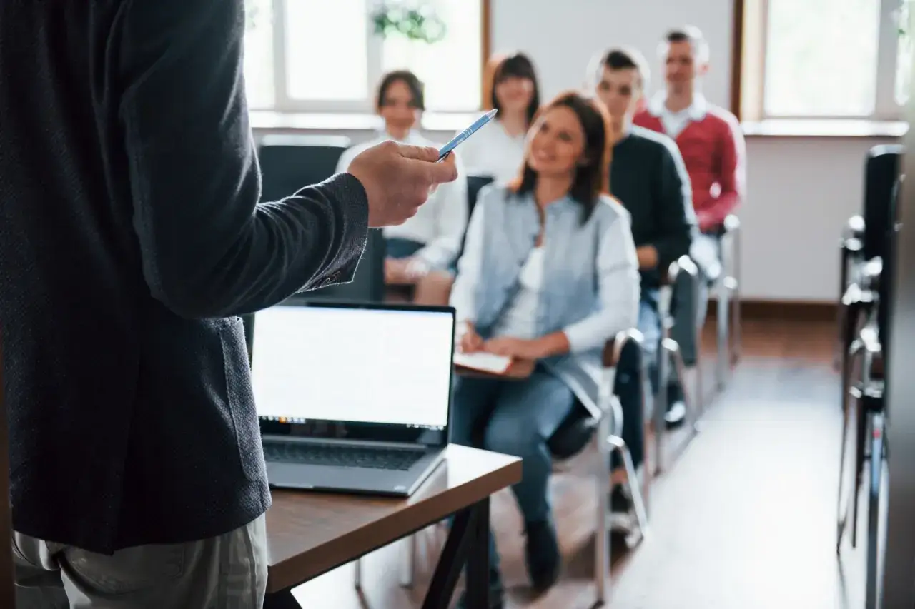 Docente con penna in mano guida un corso di scrittura. Studenti attenti siedono in aula, alcuni prendono appunti.