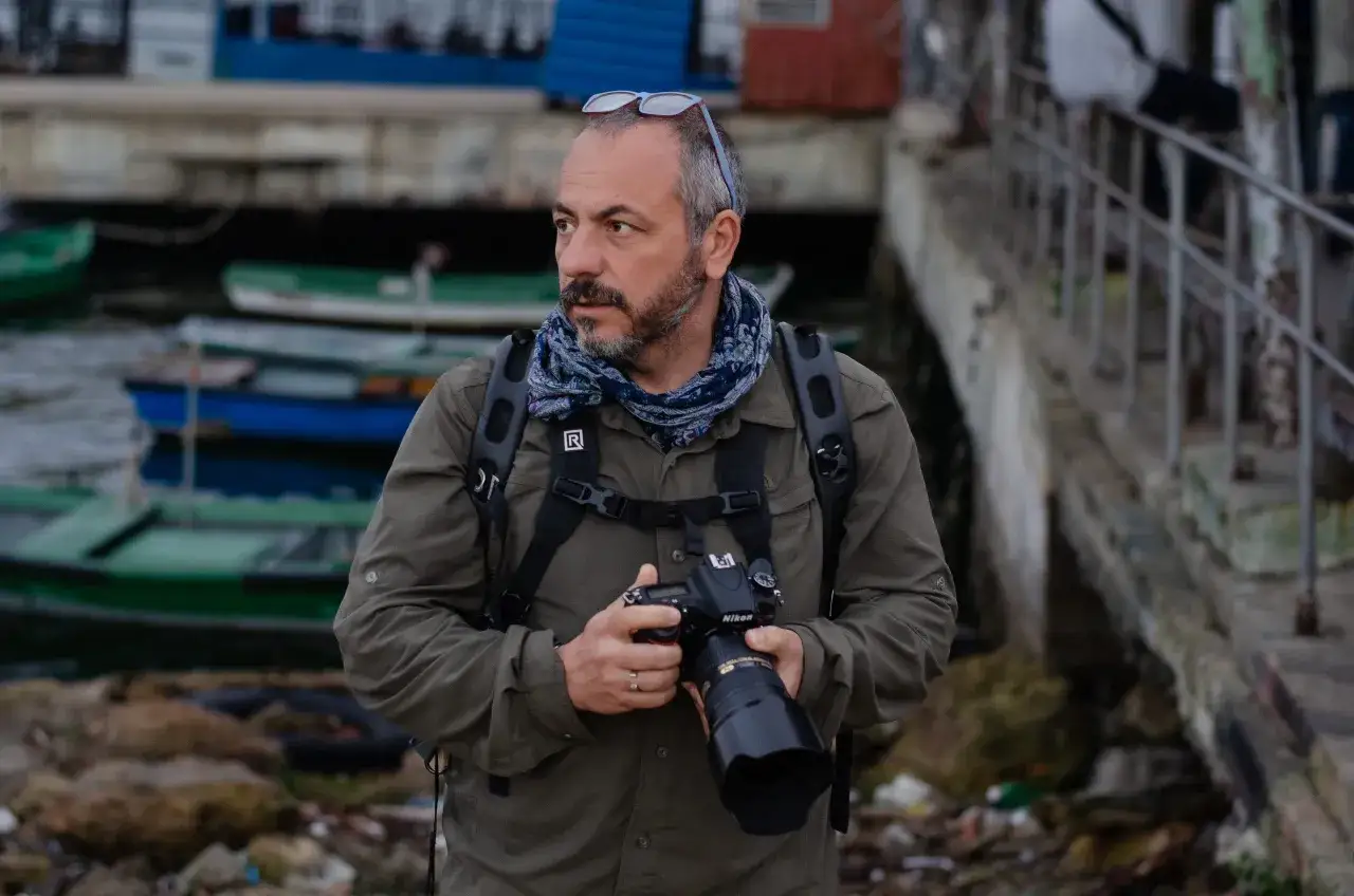 Un uomo con macchina fotografica, occhiali da sole sulla testa e sciarpa, pronto a documentare la realt&agrave;. Il suo sguardo &egrave; attento, tipico di un fotoreporter.