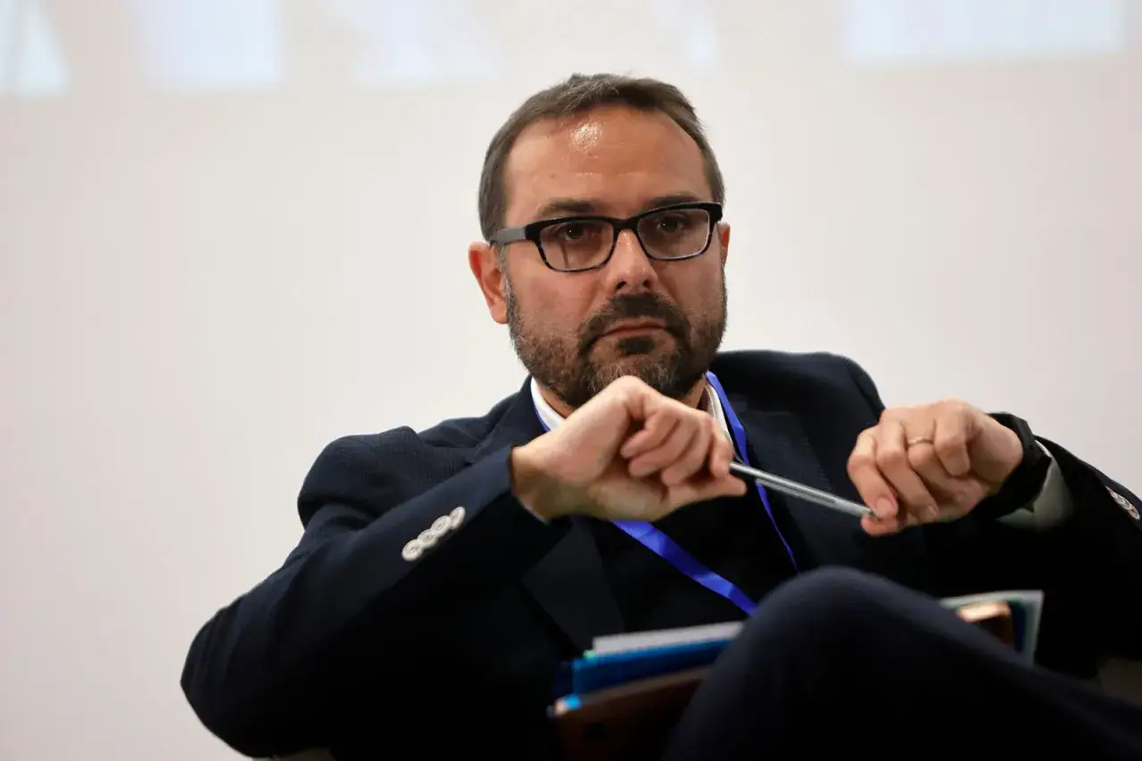 Alessandro Bollo, con occhiali e barba, tiene una penna tra le mani, pensieroso.