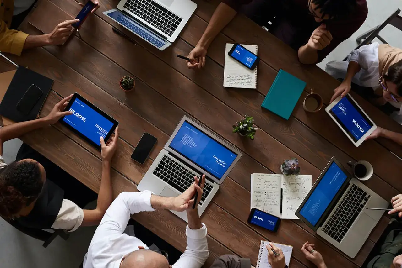 Persone attorno a un tavolo di legno, con laptop, tablet e smartphone. Il dematerializzazione significato &egrave; evidente: tutto &egrave; digitale, niente carta.