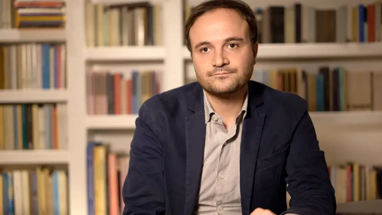 Paolo Di Paolo, con la sua aria pensierosa, &egrave; seduto davanti a una libreria piena di volumi.