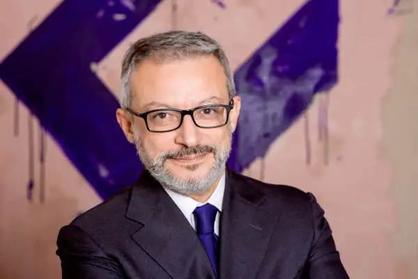 Gianluca Foglia, con occhiali e cravatta blu, sorride davanti a uno sfondo astratto viola e rosa.