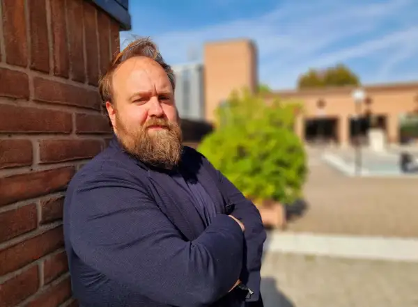 Nicolas Ballario, con barba folta e sguardo intenso, appoggiato a un muro di mattoni rossi, con un blazer blu.