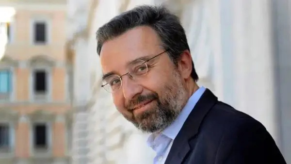 Marco Damilano, giornalista con barba e occhiali, sorride con un blazer scuro.