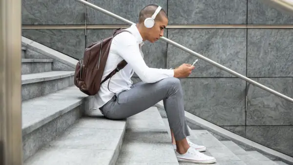 Giovane con cuffie bianche e zaino marrone seduto sulle scale, smartphone in mano, perfetto per scoprire dove ascoltare podcast.