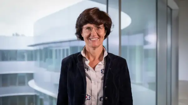 Paola Dubini, sorridente con occhiali e giacca scura, sullo sfondo di un edificio moderno con ampie vetrate.