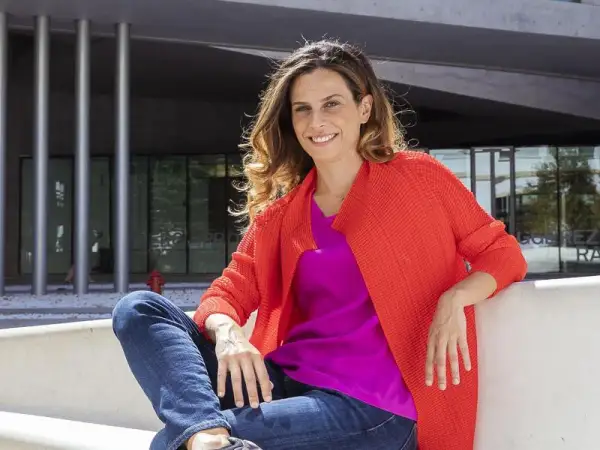 Francesca Bria sorride, seduta all'esterno con un cardigan arancione acceso e una maglia fucsia.