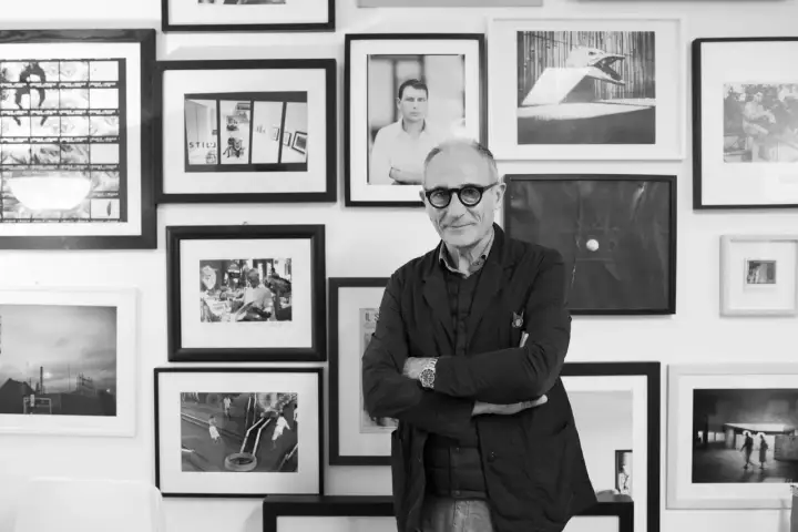 Denis Curti, circondato da opere fotografiche, sorride con le braccia conserte.